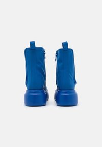 Une paire de bottines à plateforme bleu vif avec fermeture éclair à l'arrière et languette de tirage, présentée de l'arrière sur un fond blanc uni.