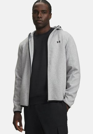 Man draagt een lichtgrijze hoodie met rits over een zwart shirt en zwarte broek, staand met één hand in de zak tegen een effen witte achtergrond.