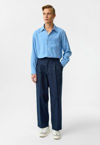 Lichtblauw button-up overhemd met een borstzak, gecombineerd met donkerblauwe wijde broek en witte sneakers met grijze accenten.