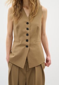 Gilet marrone su misura con scollo a V, sei bottoni neri e tasche laterali, abbinato a pantaloni larghi coordinati. Tessuto dalla texture liscia.