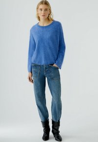 Blauwe fluffy sweater met een ronde hals, gecombineerd met lichtblauwe jeans met opgestikte zakken en zwarte enkellaarsjes.