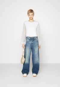 Vit topp med långa, genomskinliga ärmar och fransar; lösa blå denimjeans; krämfärgade sneakers; håller en beige väska; minimalistisk bakgrund.
