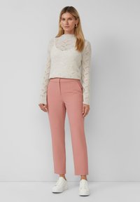 Vrouw in een crèmekleurige gebreide blouse met lange mouwen, hooggetailleerde roze broek, witte sneakers en gouden creolen, staande tegen een grijze muur.