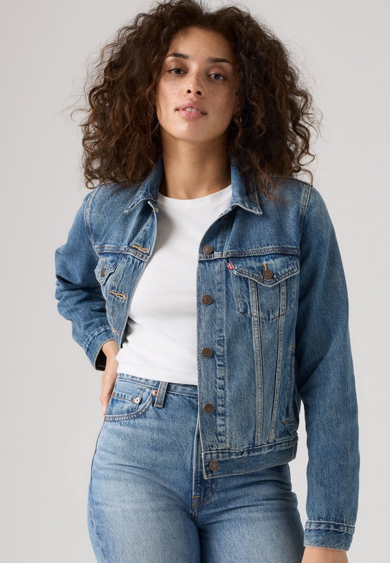 Denim jacket in light blue with a button front, chest pockets, and a collared neckline. Worn over a white T-shirt, paired with jeans.