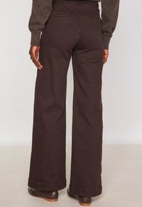 Persona con pantaloni larghi marrone scuro, scarpe marroni e un top a maniche lunghe maculato marrone, che si trova davanti a uno sfondo semplice e chiaro.