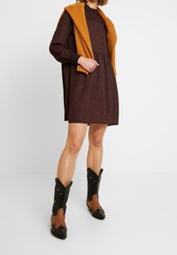 Brun mönstrad klänning med långa ärmar, kombinerad med en senapsgul tröja som hänger över axlarna. Svarta cowboystövlar med bruna accenter och stjärndetaljer.