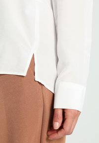 Chemise blanche à manches longues avec une texture légèrement transparente, présentant un ourlet asymétrique et un poignet blanc net. Pantalon marron texturé partiellement visible.