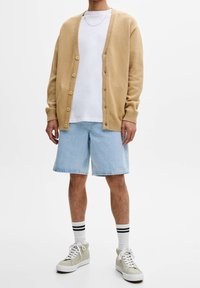 Světlé béžové kardigán s texturovaným pletením, se sedmi knoflíky, nošený přes bílé tričko, v kombinaci se světle modrými džíny a zelenými teniskami.