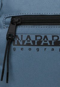 Sac à dos en tissu bleu avec une surface texturée, présentant une fermeture éclair noire et un logo imprimé. La fermeture éclair dispose d'une tirette solide.