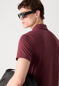 Bordový polo tričko s krátkymi rukávmi, hladká textúra a klasický golier. Nosí čierne slnečné okuliare a drží čiernu tašku s prelisovaným logom.