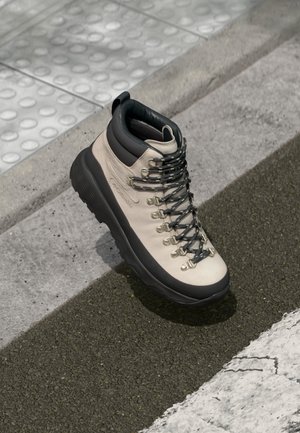 Beige hiking boot with a black padded collar, textured surface, metal eyelets, and black laces. Thick rubber sole for traction.