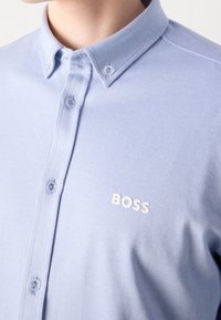 Ljusblå oxfordskjorta med texturerad väv, med en spetsig krage och en vit "BOSS"-logotyp på bröstet. Små blå knappar.