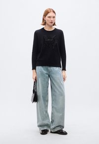 Felpa nera con logo "DKNY" in strass, abbinata a jeans a gamba larga di colore azzurro chiaro e scarpe nere. La modella tiene in mano una piccola borsa nera.