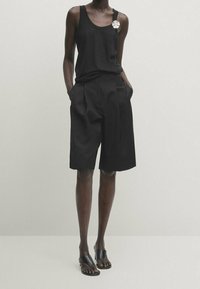 Personne portant un haut noir sans manches, un short noir ample jusqu'aux genoux, des sandales noires ouvertes aux orteils, les mains dans les poches, sur fond uni.