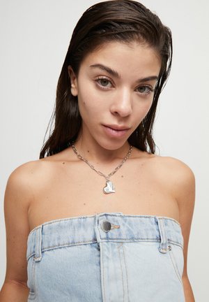 Top sin tirantes de mezclilla en azul claro con accesorios de plata y un colgante plateado en forma de corazón en una cadena. Textura suave en toda la prenda.