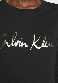 Svart sweatshirt med en centralt placerad silverlogotyp som säger "Calvin Klein" i kursiv stil. Tyget verkar mjukt och texturerat.