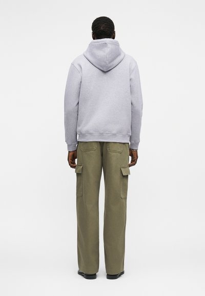 Άνδρας που φοράει ανοιχτόγριζο φούτερ με κουκούλα και χακί παντελόνι cargo, στέκεται με την πλάτη στραμμένη σε απλό λευκό φόντο.