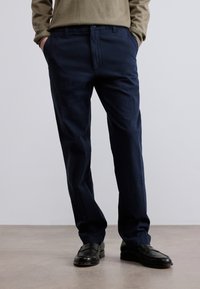 Μπλε ναυτικό chino παντελόνια από βαμβακερό μείγμα ύφασμα, με σχεδιασμό ίσιας γραμμής, θηλιές για ζώνη και δύο πλαϊνές τσέπες. Φοριούνται με μαύρα παπούτσια.