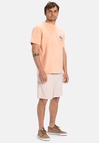 Camiseta de algodón de color naranja claro con cuello redondo y un logo en el pecho, combinada con pantalones cortos de color beige claro y zapatillas de ante marrones.