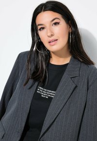 Harmaa pinstripe-puku, joka on valmistettu tekstuuri-kankaasta, päällä musta T-paita valkoisella tekstillä. Sisältää lovetun kauluksen ja suuret korvakorut.