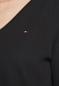 Czarny, teksturowany sweter z dekoltem w serek posiada małe, haftowane logo w kolorze czerwonym, białym i niebieskim po lewej stronie.