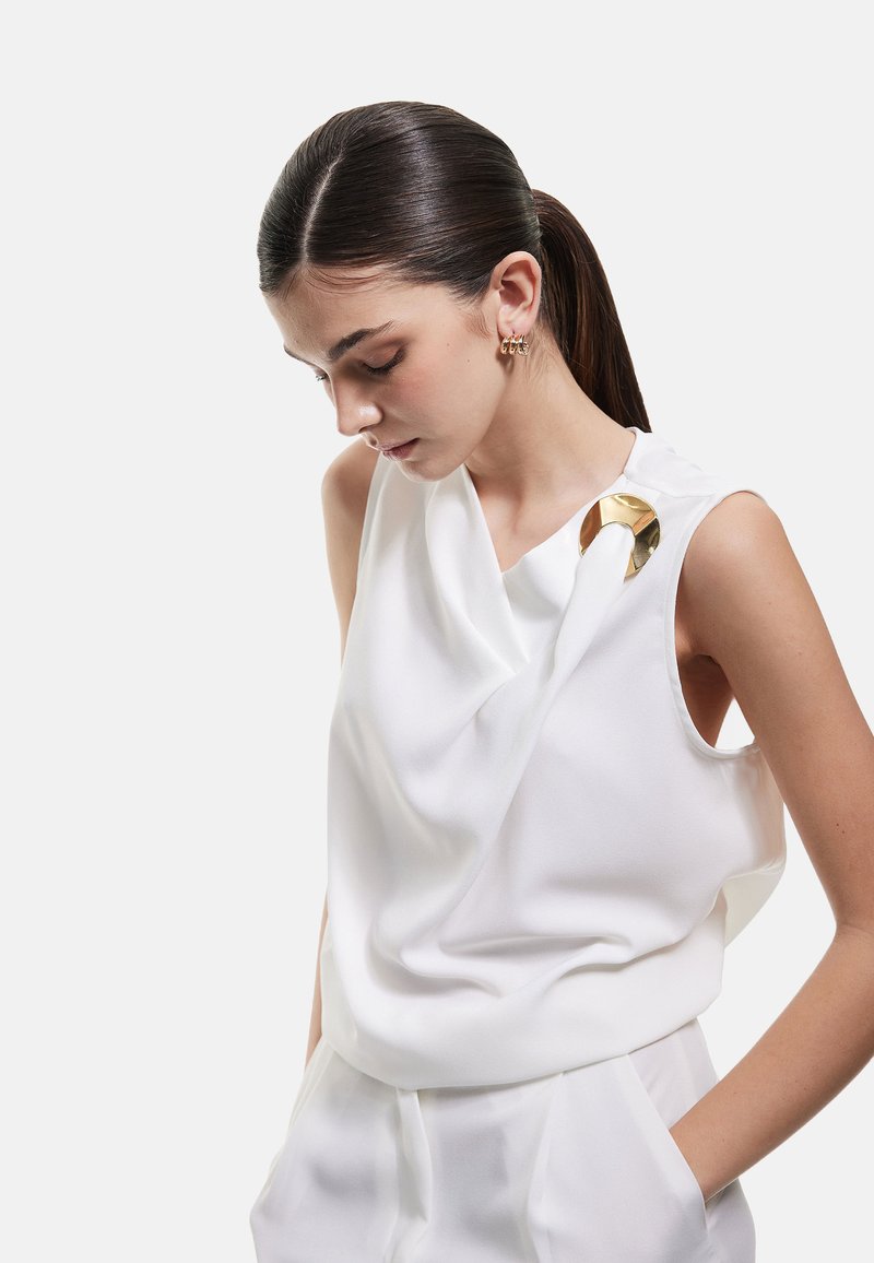 Top bianco senza maniche con design drappeggiato, texture liscia e un accento dorato sulla spalla. Abbinato a pantaloni di colore chiaro.