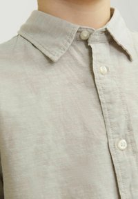 Camicia di lino grigio chiaro con colletto classico, caratterizzata da una texture sottile e chiusure a bottone. Il tessuto ha un aspetto morbido e informale.