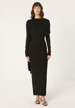 Žena stojaca v dlhej čiernej priliehavej šatke s drapovaným detailom na jednom ramene a čiernych sandáloch na opätku s remienkami na jednofarebnom pozadí.