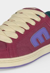 Gros plan d'une basket bordeaux avec de gros lacets crème, un patch logo violet et une semelle crème avec un liseré violet.