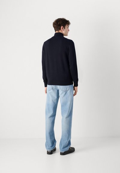 Sudadera azul marino con cuello alto, puños ajustados, combinada con jeans de corte relajado en azul claro y zapatos negros, vista desde la parte trasera.