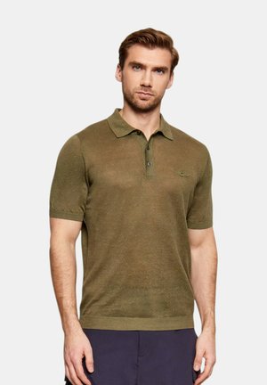 Uomo adulto che indossa una polo aderente verde oliva a maniche corte con due bottoni, in piedi davanti a uno sfondo chiaro e uniforme.