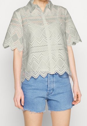 Lysegrå blondebluse med geometriske mønstre og bølget kant sammen med frynset denimshorts på en stående person.