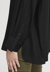Maglietta nera a maniche lunghe con dettagli elastici sui polsini, tessuto testurizzato e accentata da bottoni. Abbinata a pantaloni verde oliva, mostrando un fit rilassato.