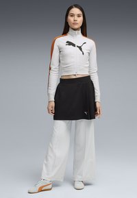 Witte cropped jas met oranje accenten en zwart Puma-logo, gecombineerd met een zwarte rok en witte wijde broekspijpen. Grijze schoenen met oranje detail.