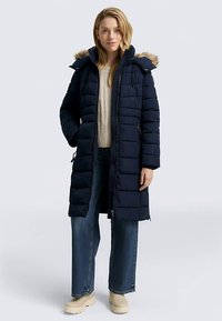 Doudoune marine avec capuche bordée de fourrure, fermeture éclair, texture matelassée et poches latérales. Associée à un pull beige et un jean bleu.