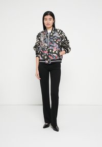 Chaqueta bomber de tela negra con un diseño multicolor que incluye animales y estrellas; cuenta con puños y cinturilla a rayas. Pantalones negros puestos.