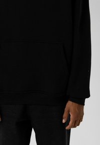 Zwart sweatshirt met een voorzak, gemaakt van zachte stof. Voorzien van lange mouwen en een comfortabele pasvorm. Gecombineerd met een donkere spijkerbroek.