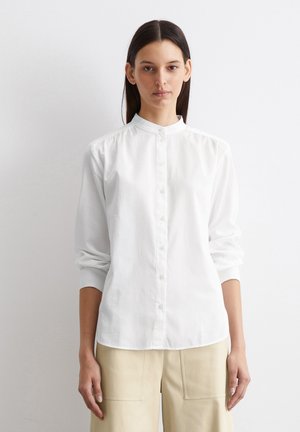 Biała koszula zapinana na guziki z kołnierzykiem stójkowym, długimi rękawami i marszczeniami na ramionach, wykonana z gładkiego, lekkiego materiału.