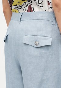 Ανοιχτό μπλε παντελόνι με φερμουάρ, με τσέπες με καπάκια, το ένα κουμπωμένο, φορεμένο με πουκάμισο με σχέδιο που είναι μπηγμένο στη μέση.