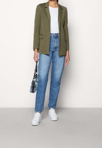 Blazer en lin vert olive, t-shirt blanc, jean taille haute bleu clair, baskets blanches, tenant un sac à main noir et turquoise à motifs.