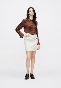 Chemise en satin marron à manches longues avec boutons, assortie à une mini-jupe en cuir blanc dotée de poches avant et de boutons dorés.