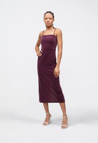 Femme debout, main sur la hanche, portant une robe midi prune sans manches et des sandales blanches à talons hauts, sur un fond uni.