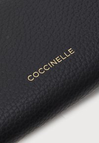 Black textured leather wallet with embossed "Coccinelle" logo in gold. Features a pebbled surface for added durability and grip.