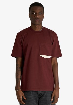 Giovane uomo che indossa una t-shirt bordeaux a maniche corte con taschino frontale e jeans neri, in piedi su uno sfondo bianco neutro.
