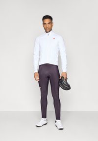 Weiße Radjacke mit Reißverschluss, dunkelviolette Radhosen und weiße Radschuhe. Die Person hält einen schwarzen Helm.