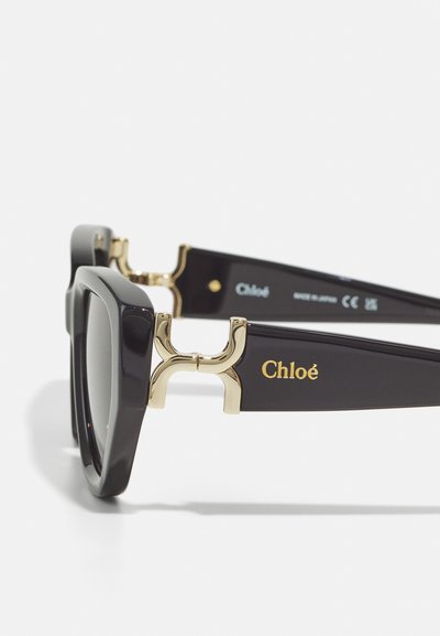 Black oversized sunglasses with square frames, gold accents at the temples, and "Chloé" logo embossed in gold on the side.