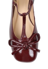Bordowe lakierowane buty na wysokim obcasie z designem T-strap, zdobione dużą kokardą i węzłem na czubku, o gładkiej teksturze i zaokrąglonym kształcie.