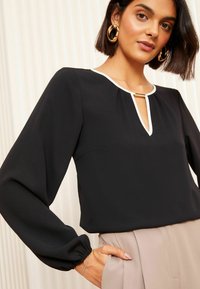 Blusa nera a maniche lunghe con scollatura bianca e dettaglio a chiave, dotata di polsini elastici e una texture liscia.