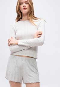 Νέα γυναίκα με μακριά μαλλιά στέκεται με σταυρωμένα τα χέρια, φοράει γκρι ανοιχτόχρωμο υφασμάτινο μακρυμάνικο τοπ και ταυτόχρονες σορτς με βολάν στα άκρα.