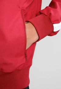 Chaqueta bomber roja con acabado satinado suave, puños acanalados y un bolsillo lateral con cremallera. Presenta un color sólido sin patrones ni acentos.